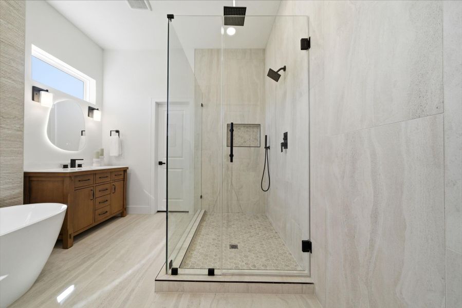 This modern bathroom features a spacious glass-enclosed shower with sleek fixtures, a freestanding bathtub, and a wooden vanity with dual sinks. The neutral tones and natural light enhance its elegant and airy atmosphere.