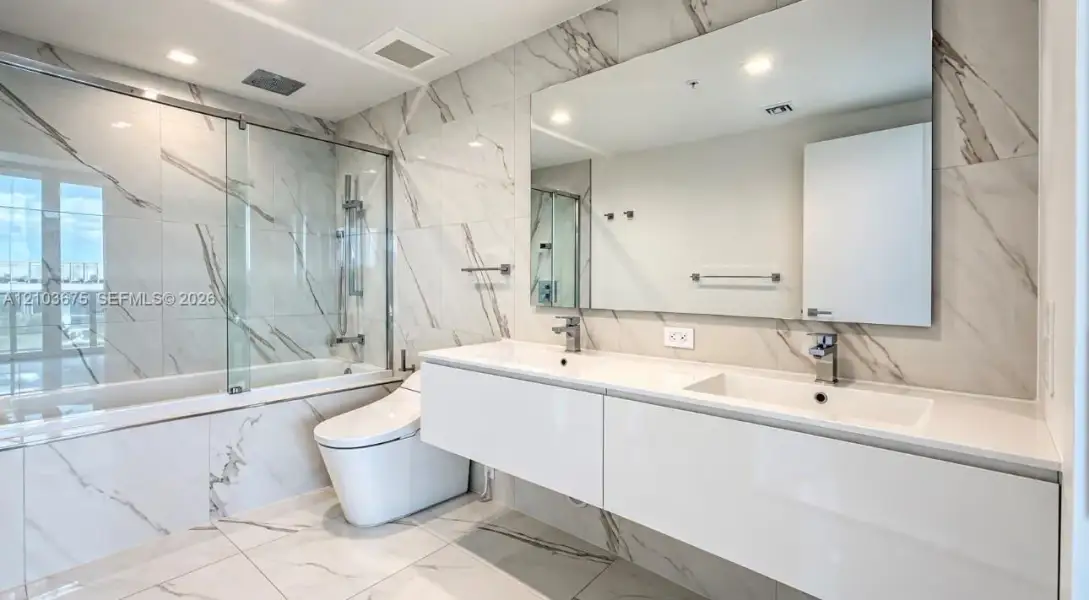 Floating dual-sink vanity with a solid surface countertop, large wall-mounted mirror, floor-to-ceiling stone-patterned tile, and a glass-enclosed shower and tub combination Floating dual-sink vanity with a solid surface countertop, large wall-mounted mirror, floor-to-ceiling stone-patterned tile, and a glass-enclosed shower and tub combination