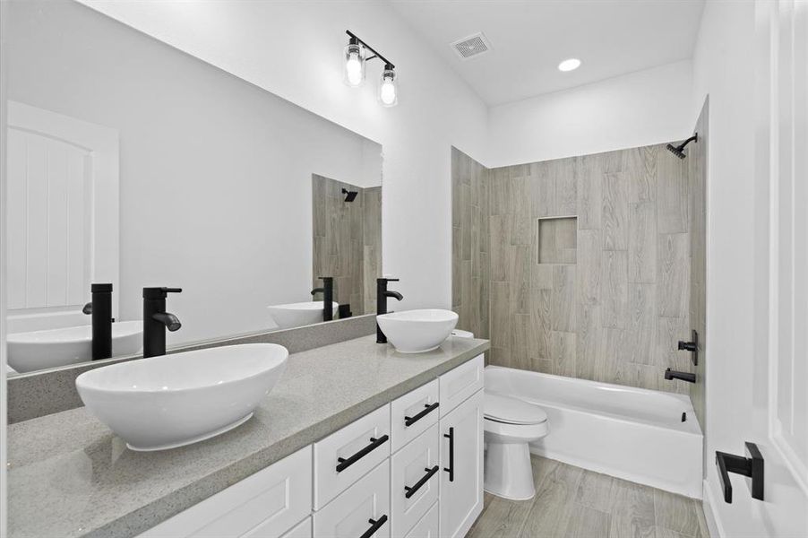 The bathroom features a modern design with dual vessel sinks, sleek black fixtures, a tiled shower-tub combo, and wood-look tile flooring. There's ample storage with white cabinetry and a well-lit mirror area.