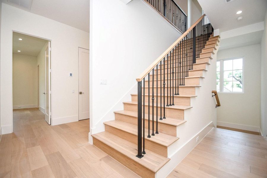 This photo features a bright, modern entryway with light wood flooring and a staircase with wooden steps and black metal railings. The area is well-lit with recessed lighting and a window, offering a welcoming and spacious feel.