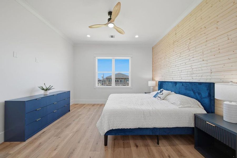 This spacious bedroom features a modern design with a wooden accent wall and light hardwood flooring. It includes a stylish blue bed, matching dresser, and nightstands. A large window lets in natural light, and a ceiling fan adds comfort.