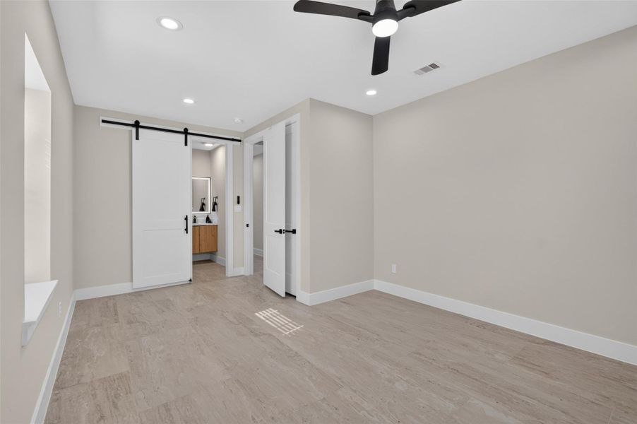 Spacious room with elegant wood flooring, a modern ceiling fan, and a sleek barn door leading to a bathroom, offering a stylish and functional living space.