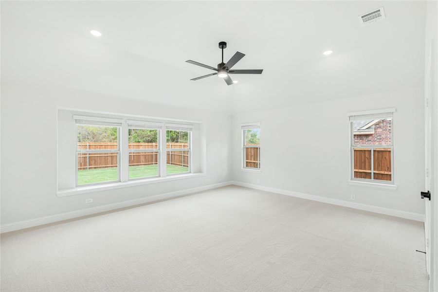 Empty bedroom with carpet flooring, ceiling fan, and multiple windows overlooking a fenced backyard