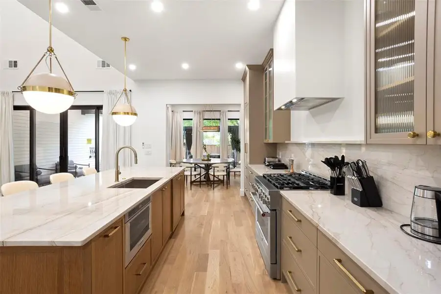 Kitchen featuring light wood-finish flooring, a large central island with an integrated sink and microwave, pendant lighting, stone countertops with a matching backsplash, and stainless steel appliances