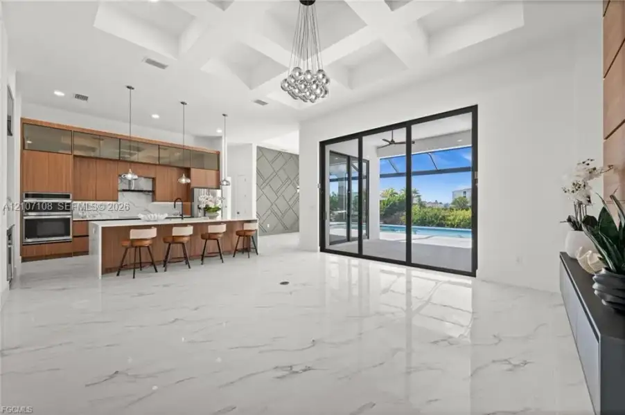 Coffered ceilings with recessed lighting, large-format tile flooring, wood-finish cabinetry, built-in stainless steel double ovens, and multi-panel sliding glass doors