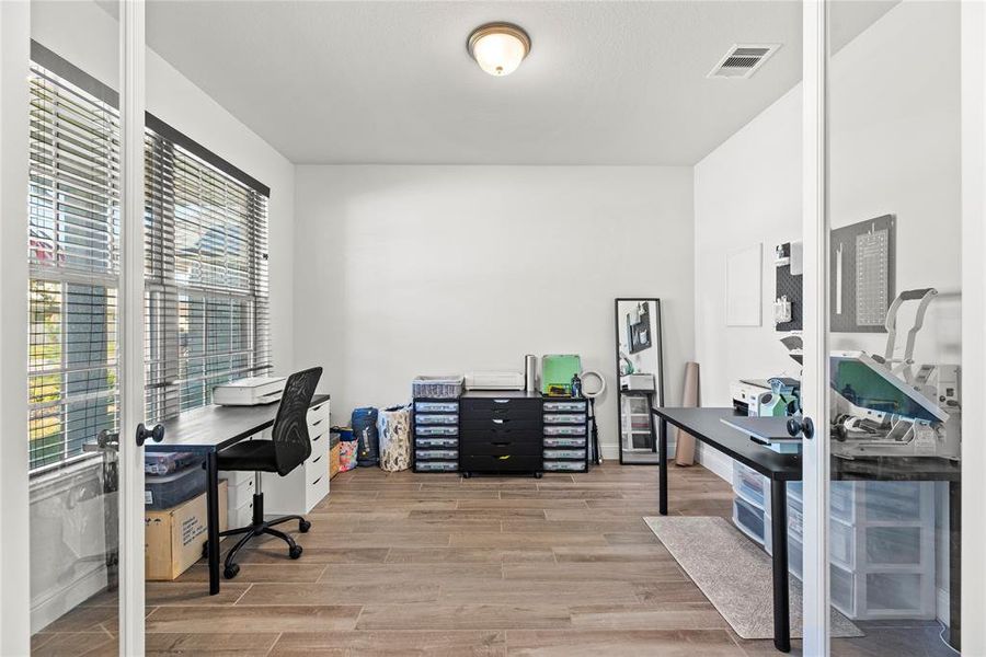 This is a well-lit, versatile room that could serve as a home office or craft space, featuring large windows with blinds, neutral walls, and wood-look flooring. It includes glass French doors that enhance the open feel of the space.