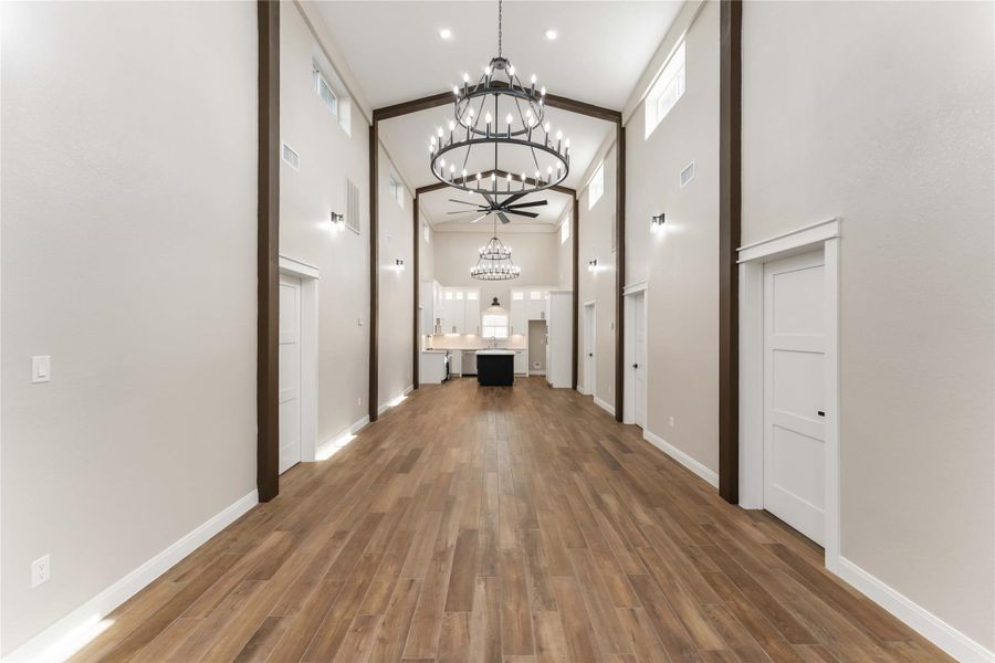 This photo showcases a modern, open-concept living area with high ceilings and elegant chandeliers. The space features beautiful tile wood-style flooring and ample natural light, providing a warm and inviting atmosphere. The view extends to a kitchen area with a central island, perfect for entertaining.
