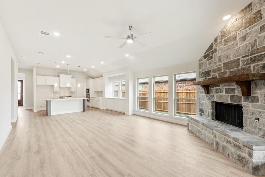 Open living area with stone fireplace, light wood floors, and white kitchen in background