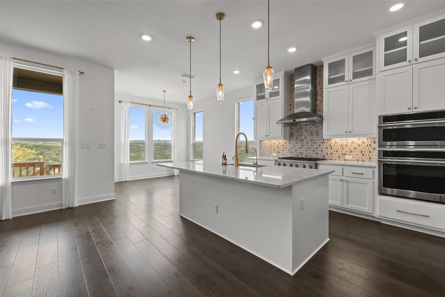 Kitchen featuring backsplash, a center island with sink, a sink, double oven, and wall chimney exhaust hood