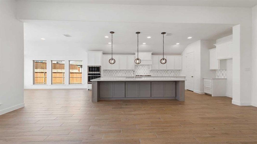 Kitchen featuring light wood-type flooring, backsplash, pendant lighting, white cabinets, and a kitchen island with sink