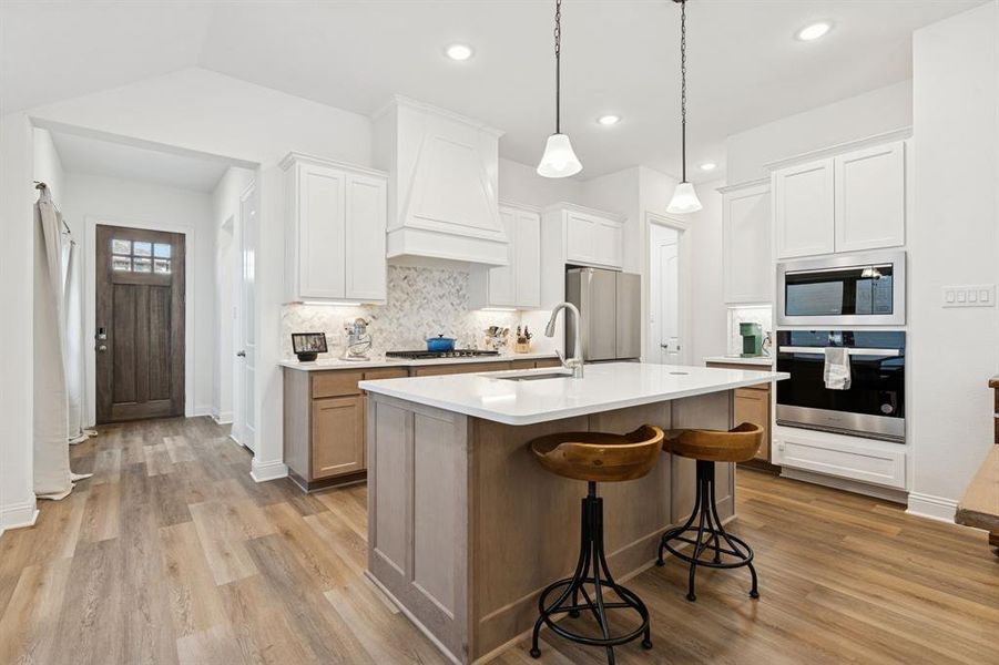 Kitchen with decorative backsplash, stainless steel appliances, white cabinets, an island with sink, and light wood-style flooring Kitchen with decorative backsplash, stainless steel appliances, white cabinets, an island with sink, and light wood-style flooring