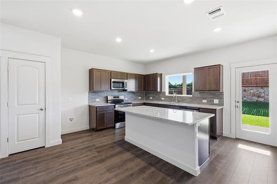 Kitchen featuring an expansive island with a light-colored countertop, wood-finish flooring, dark wood cabinetry, a subway tile backsplash, and stainless steel appliances