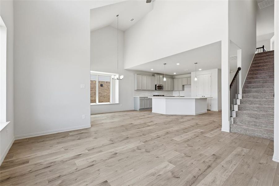 Open-concept living space featuring vaulted ceilings, light wood-finish flooring, a kitchen island with pendant lighting, and an adjacent carpeted staircase with a dark banister
