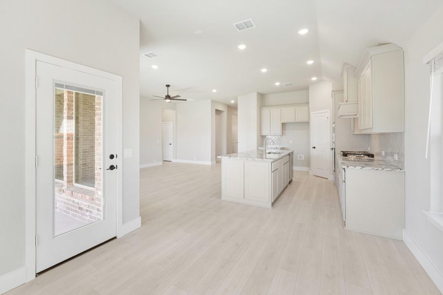 Open kitchen with white cabinets, center island, granite countertops, and light wood floors open to living area with ceiling fan.