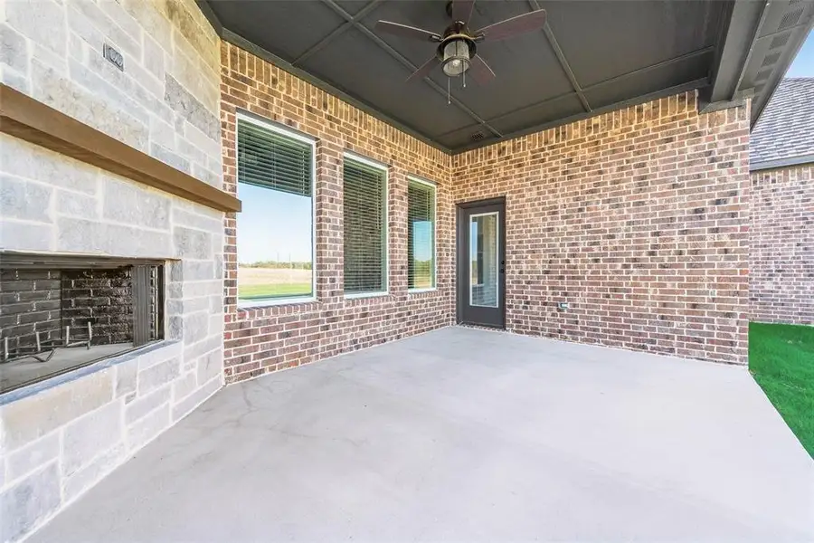 Expansive outdoor patio featuring an integrated stone fireplace with a wood mantle, concrete flooring, and a ceiling fan