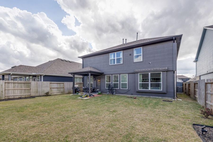 Spacious backyard with a two-story, gray house featuring large windows and a covered patio. It's enclosed by a wooden fence, providing privacy and a sizable lawn area. Perfect for outdoor activities and relaxation.