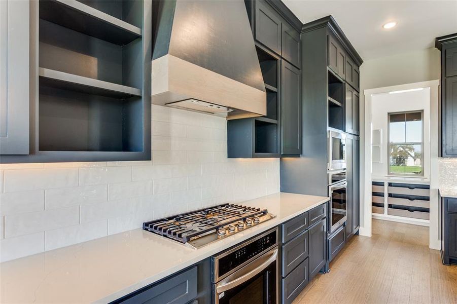 Kitchen with custom exhaust hood, open shelves, stainless steel appliances, tasteful backsplash, and light stone countertops Kitchen with custom exhaust hood, open shelves, stainless steel appliances, tasteful backsplash, and light stone countertops