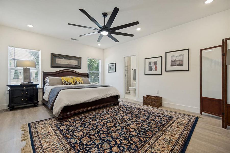 Spacious bedroom featuring light wood-finish flooring, a modern black ceiling fan, recessed lighting, and two windows providing natural light Spacious bedroom featuring light wood-finish flooring, a modern black ceiling fan, recessed lighting, and two windows providing natural light