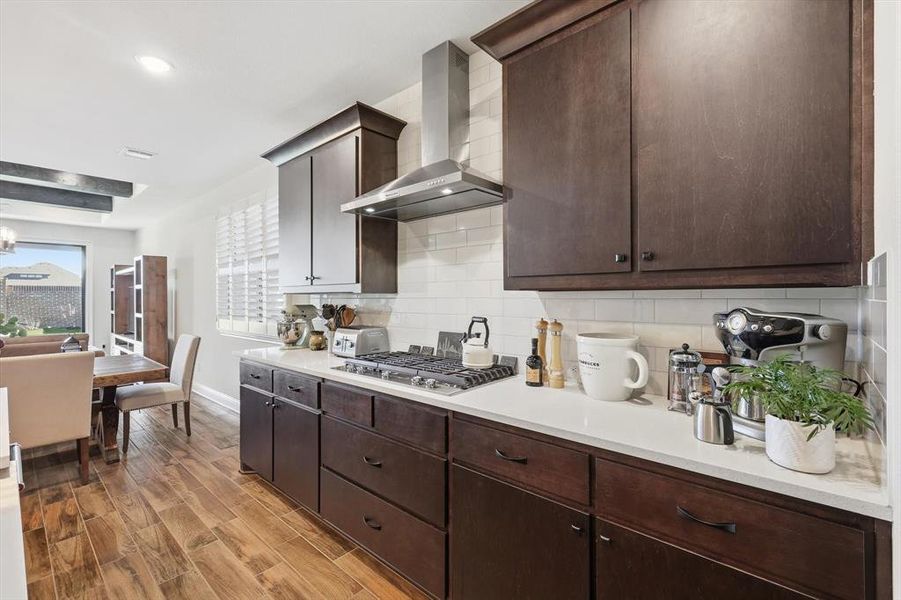Kitchen with dark brown cabinets, wall chimney exhaust hood, wood tiled floors, backsplash, and stainless steel gas cooktop