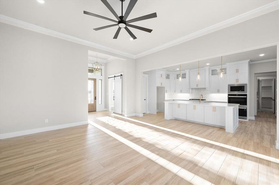 Unfurnished living room featuring ceiling fan with notable chandelier, a barn door, light hardwood / wood-style flooring, and ornamental molding