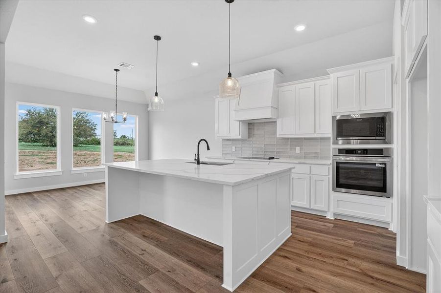 Open-concept kitchen featuring a white center island with a sink, white cabinetry, integrated stainless steel appliances, a light-toned tile backsplash, and wood-finish flooring