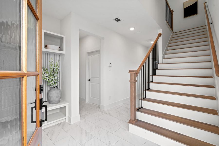 A bright entryway with a wooden front door, leading to a staircase with wooden steps and a metal railing. The space features white walls, tiled floors, and modern decor, creating a welcoming and contemporary atmosphere.