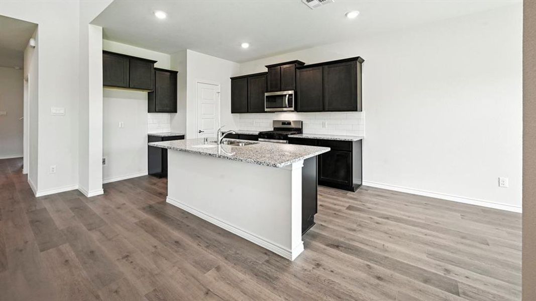 Kitchen featuring stainless steel appliances, light stone counters, tasteful backsplash, an island with sink, and light wood-style flooring