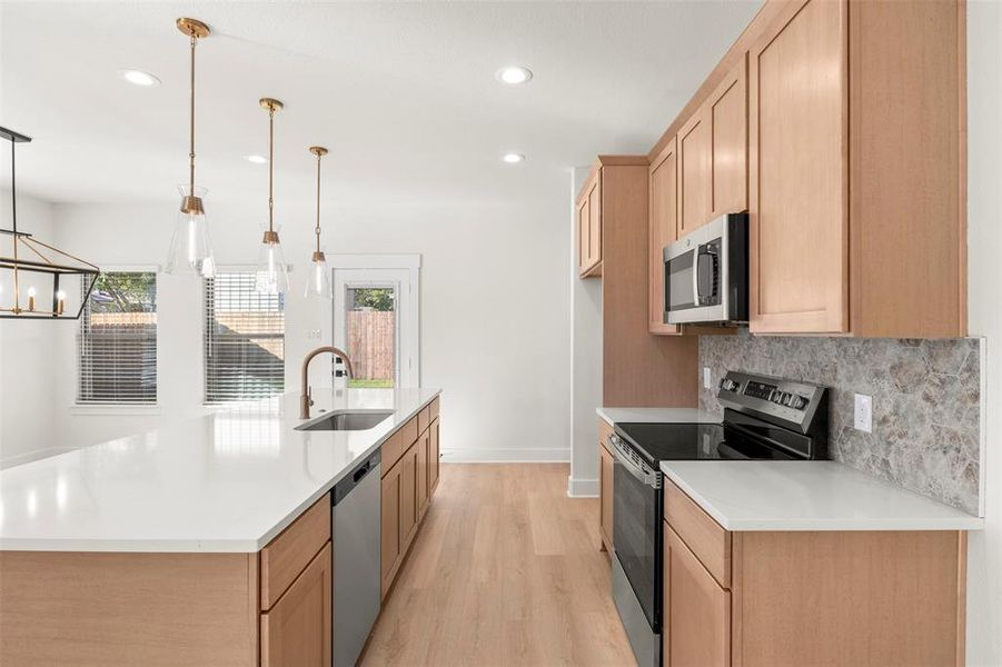 Kitchen featuring appliances with stainless steel finishes, light wood-style flooring, light brown cabinetry, decorative light fixtures, and an island with sink