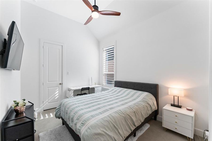 This room features a vaulted ceiling with a ceiling fan, a window with plantation shutters, and light-colored carpeting