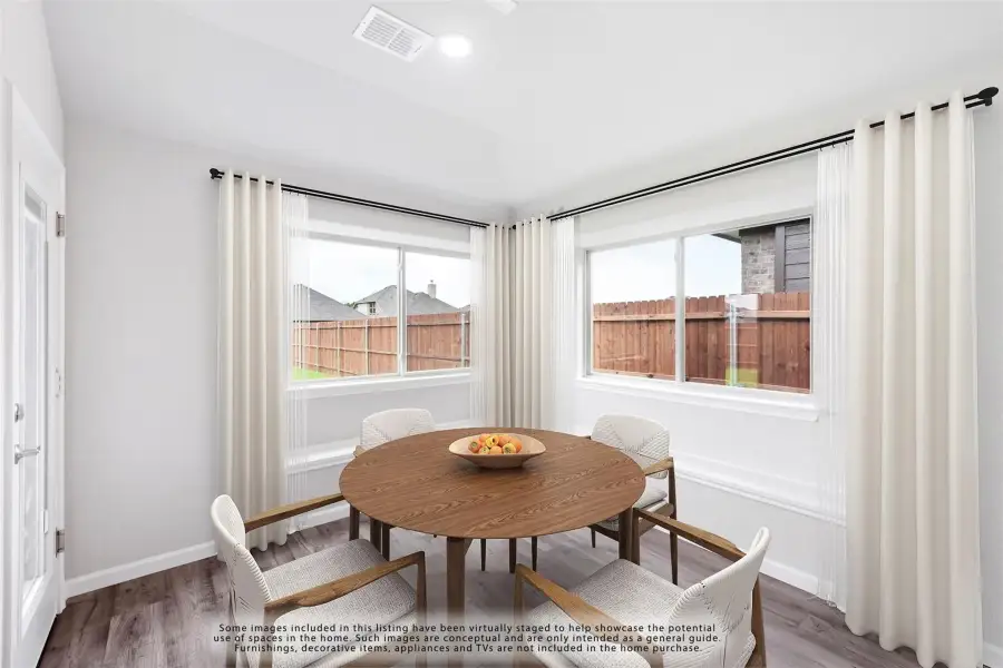 Dining area with round wood table, four chairs, hardwood floors, and two large windows with white curtains