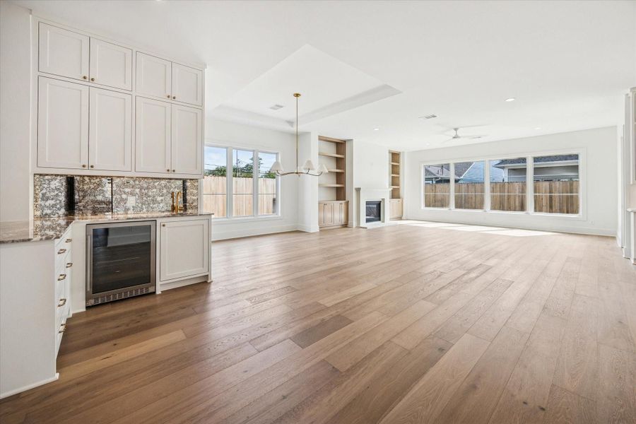Open concept living and dining space anchored by a custom wet bar with wine refrigerator, expansive engineered hardwood flooring, built in shelving, and walls of windows overlooking the backyard, seamlessly connecting the kitchen, fireplace focal point, and entertaining areas in this Harvard Homes design.