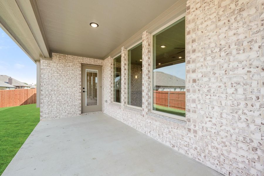 Covered back patio with concrete floor, white brick exterior, glass door, and fenced backyard with green lawn