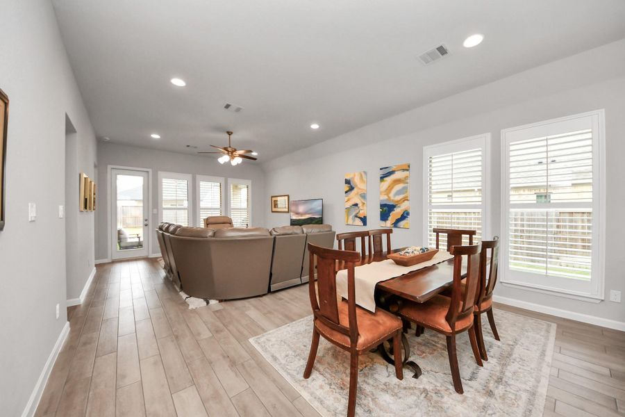 This spacious open-plan living and dining area features hardwood floors, recessed lighting, and large windows with plantation shutters for natural light. The room is tastefully decorated with contemporary artwork and offers a cozy seating area perfect for relaxation.