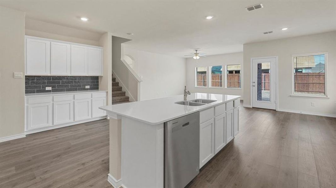 Open-concept living area featuring wood-finish flooring, a kitchen island with dual sinks and dishwasher, white cabinetry with dark subway tile backsplash, and multiple windows for natural light