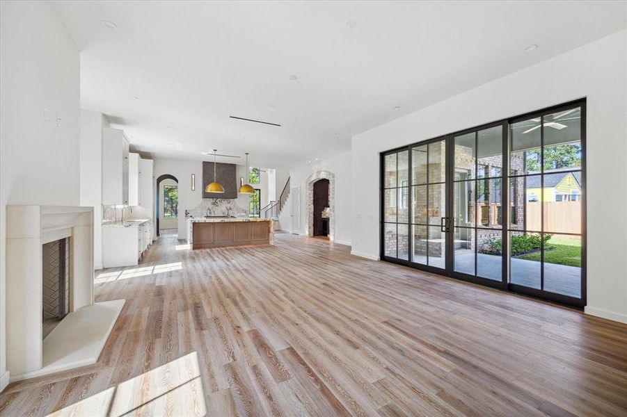 A spacious open-plan living area featuring large sliding glass doors, a gas fireplace, and a seamless transition into the kitchen, all illuminated by natural light and high ceilings and European oak wood flooring.