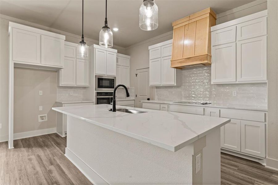 Kitchen featuring a central island with an undermount sink, white cabinetry, a built-in microwave, wood-finish flooring, and a decorative wood range hood Kitchen featuring a central island with an undermount sink, white cabinetry, a built-in microwave, wood-finish flooring, and a decorative wood range hood
