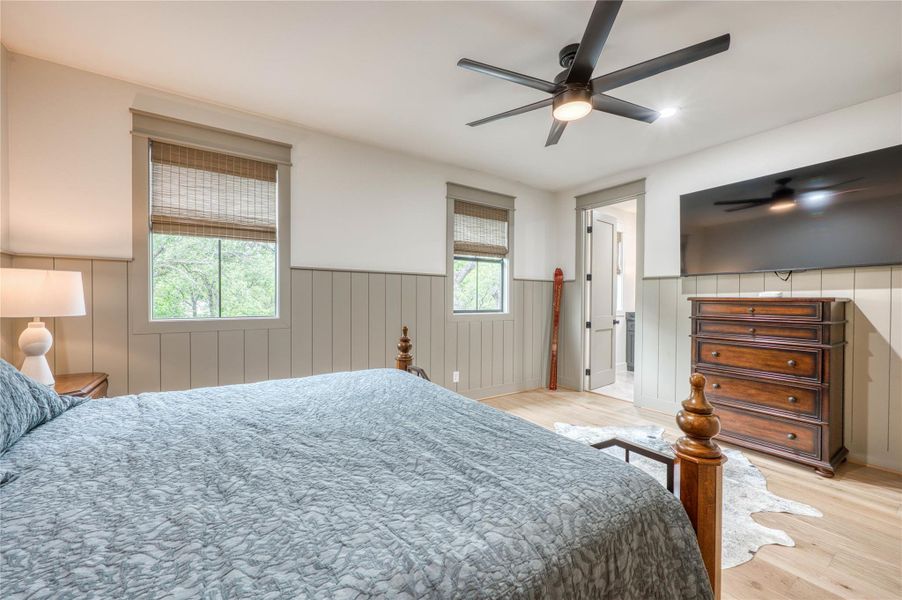 Bedroom featuring wood-finish flooring, two windows with woven shades, a ceiling fan with integrated lighting, and board and batten wainscoting