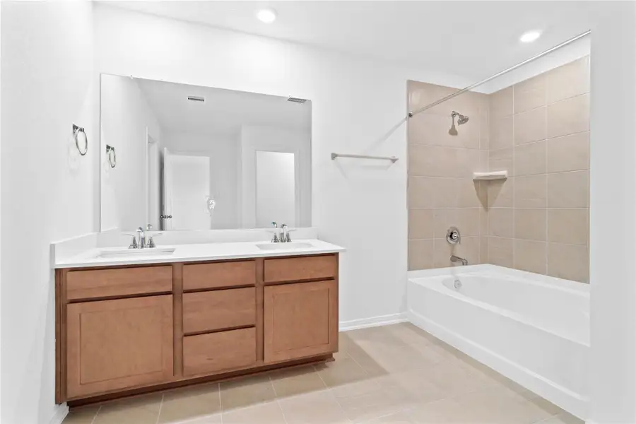 The primary bathroom offers a spacious tub/shower combination with a tiled surround, stained cabinetry topped with light countertops, dual sinks, high ceilings, neutral paint, a large mirror, and sleek, modern finishes for a polished look.