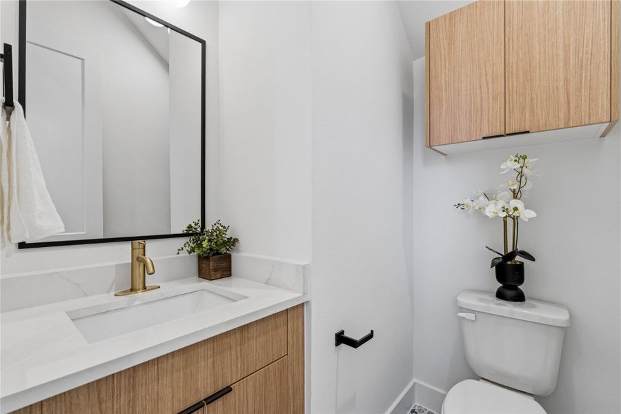 A sophisticated bath featuring a light wood vanity topped with white stone and a brushed gold faucet. Black-framed accents and patterned tiling add a modern, high-contrast touch to the compact space.