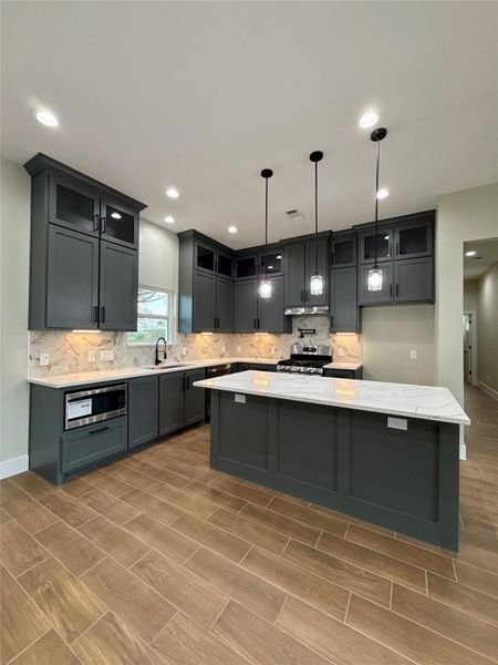 This kitchen features modern dark cabinetry with stylish marble countertops and backsplash. It includes a central island with pendant lighting, stainless steel appliances, and wood-look tile flooring, creating a sleek and inviting space.