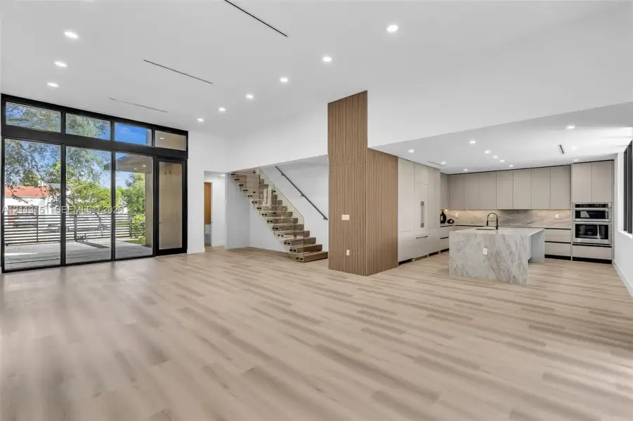 Open-concept interior featuring wood-finish flooring, floor-to-ceiling glass paneling, a central island with stone-finish surfaces, custom flat-panel cabinetry, and a floating staircase with glass railings Open-concept interior featuring wood-finish flooring, floor-to-ceiling glass paneling, a central island with stone-finish surfaces, custom flat-panel cabinetry, and a floating staircase with glass railings