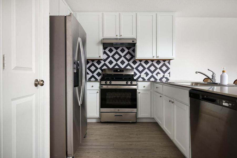 Kitchen featuring appliances with stainless steel finishes, white cabinets, backsplash, wood finished floors, and under cabinet range hood Kitchen featuring appliances with stainless steel finishes, white cabinets, backsplash, wood finished floors, and under cabinet range hood