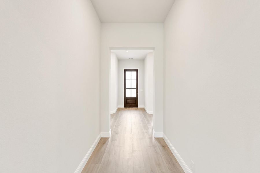 Narrow hallway with light wood floors, white walls, and dark wood front door with glass panels