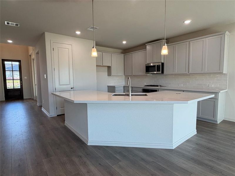 Modern kitchen featuring a large island with an undermount sink, pendant lighting, stainless steel appliances, light gray cabinetry, and wood-finish flooring Modern kitchen featuring a large island with an undermount sink, pendant lighting, stainless steel appliances, light gray cabinetry, and wood-finish flooring