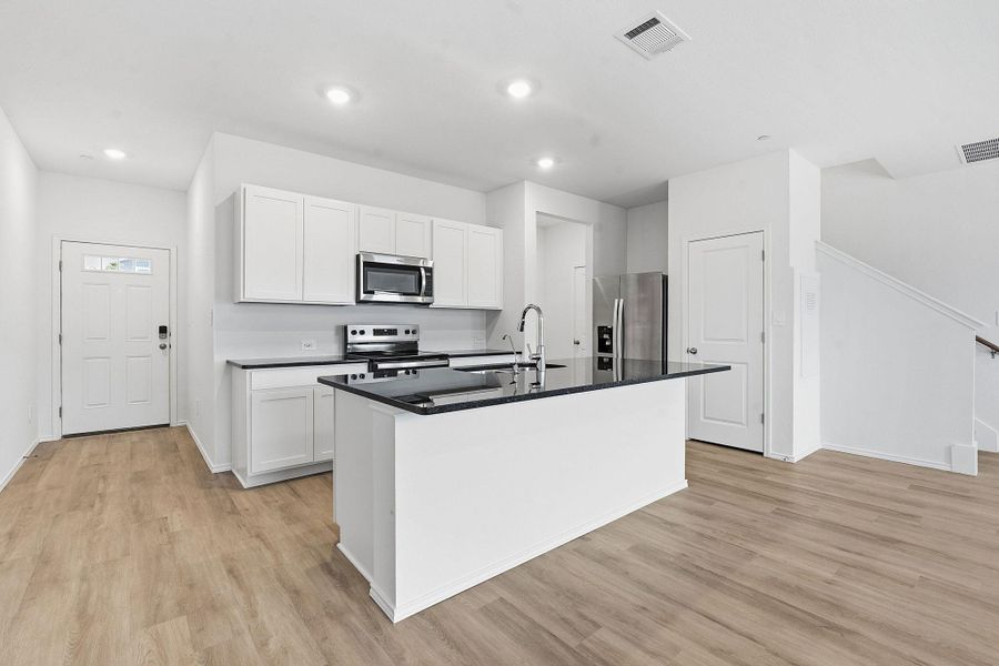Modern kitchen featuring white cabinetry, dark countertops, a central island with a sink, stainless steel appliances, and wood-finish flooring Modern kitchen featuring white cabinetry, dark countertops, a central island with a sink, stainless steel appliances, and wood-finish flooring