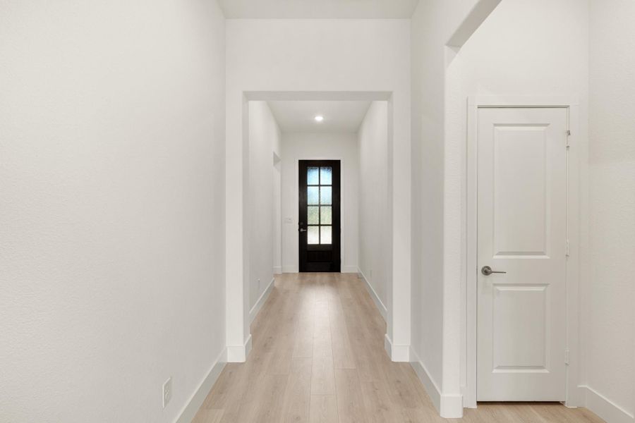 Bright hallway with light wood floors, white walls, and black glass-panel front door at the end
