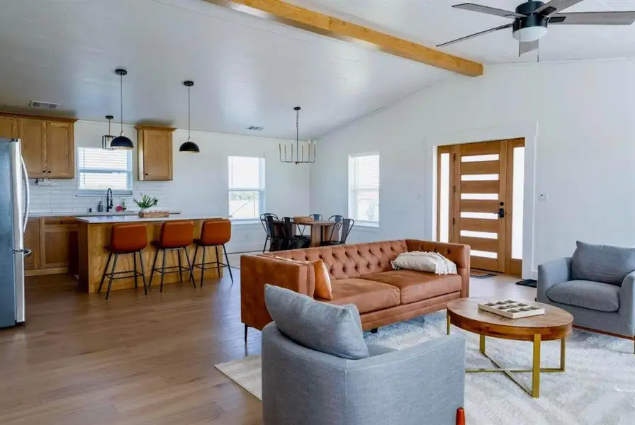 Open concept living space featuring vaulted ceilings with exposed wood beams, wood-finish flooring, a contemporary ceiling fan, and a modern wooden entry door with frosted glass panels
