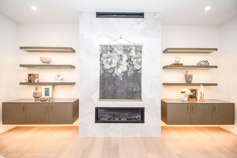 This photo showcases a modern living room with a sleek, built-in fireplace at the center, flanked by stylish floating shelves and cabinets. The light wood flooring and soft lighting create a warm, inviting atmosphere, perfect for both relaxation and entertaining.