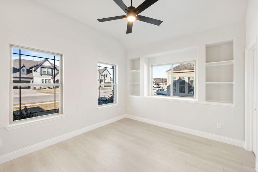 Empty room with white walls, built-in shelves, ceiling fan, and three windows overlooking a neighborhood