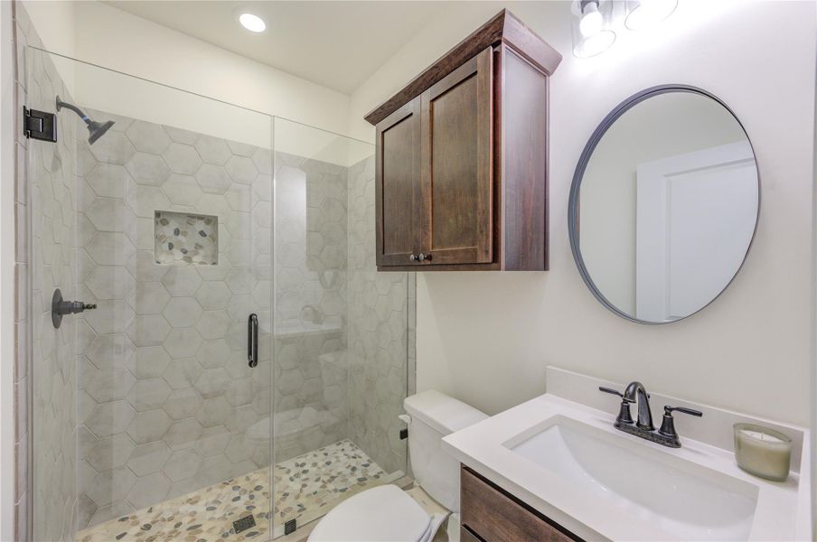 Modern secondary bathroom with a glass-enclosed shower featuring hexagonal tiles and a pebble floor. There's a sleek vanity with a round mirror and dark wood cabinetry, creating a stylish and functional space.